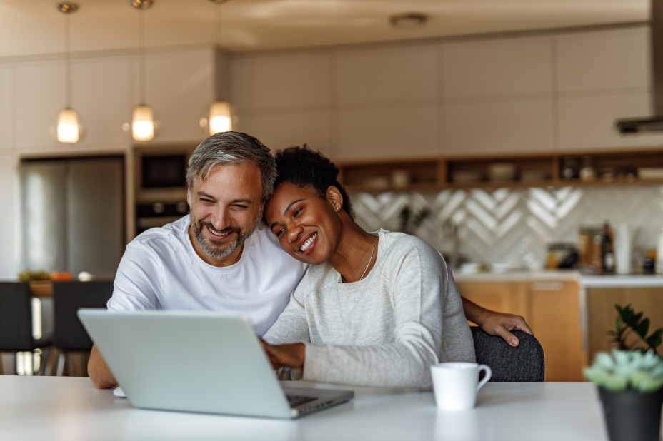 image of a Couple (woman leaning her head on man's shoulder) sitting at a table looking at a laptop smiling. 